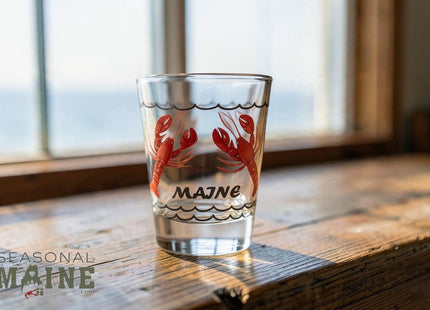 Maine Shot Glass - Lobsters — small empty souvenir shot glass in a cottage setting, Seasonal Maine logo