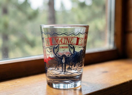 Maine Shot Glass - Lobster / Moose — small empty souvenir shot glass in a cottage setting, Seasonal Maine logo