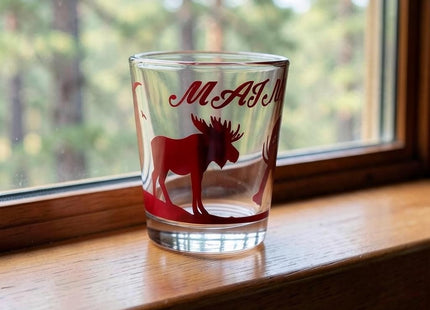 Maine Shot Glass - Red Moose — small empty souvenir shot glass in a cottage setting, Seasonal Maine logo