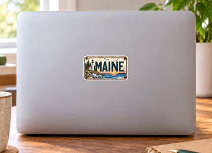 Maine Lighthouse Sunset Vintage License Plate Vinyl Sticker – 3 Inch shown on a laptop in a styled setting, with Seasonal Maine logo. Includes sticker only.