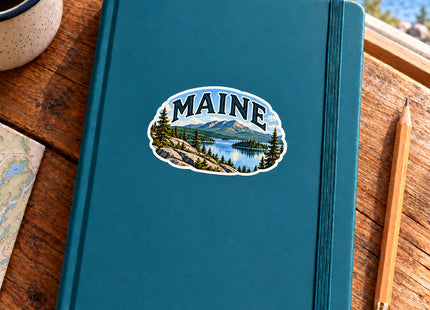 Maine Mountain Lake Vinyl Sticker – 3 Inch shown on a notebook in a styled setting, with Seasonal Maine logo. Includes sticker only.