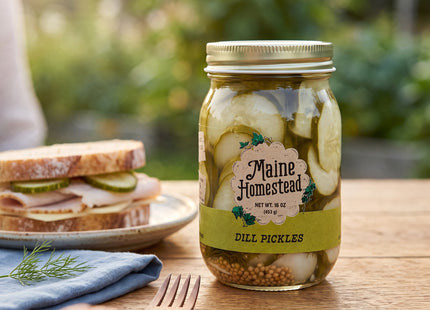 Maine Homestead Dill Pickle Slices jar beside a sandwich on a rustic picnic table; Seasonal Maine logo watermark