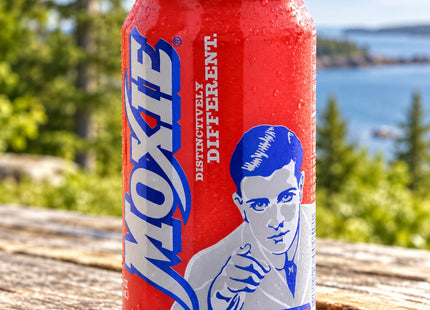 Original Moxie12fl oz can on a sunlit Maine porch table, with Seasonal Maine logo. Variety and quantity depend on selection.